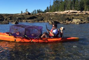 Tony Pellegrini sits in a kayak with his medium-sized lab dog, Sadie, and some lobster traps. The boat is orange and there are trees on the shoreline in the background. 