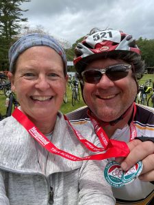 Tony Pellegrini and wife, Dr. Joan Pellegrini, are in a selfie after a long bike ride. Both are smiling. Tony is holding the medals that are around his and Joan's necks. 