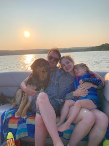 The Wagner family sits on a boat. In the background is the water of the lake at sunset. From left to right, seated is Roger, the family dog, Stephen Wagner, Cayla Wagner, and Greta Wagner, their daughter. 