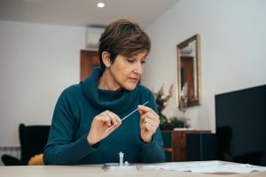 Senior woman sitting at home, studying the instructions for a self test for COVID-19 with Antigen kit on hand. Holds a nasal swab for possible infection of Coronavirus. 