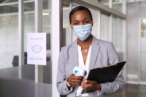 African business woman ready to use infrared forehead thermometer to check body temperature for virus symptoms before entering the office, epidemic virus outbreak. Portrait of black businesswoman wearing surgical mask holding thermometer and clipboard. Covid flu screening concept, no mask no entry sign.