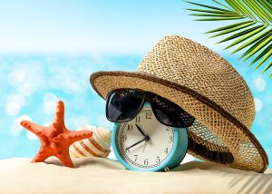 Summertime vacation concept. Time to relax. Last minute deals. Alarm clock with straw hat, starfish on the sand beach and sea background.