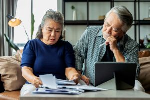 Serious stressed asian senior old couple worried about documents, retired family looking at tablet and paperwork, sitting on couch in home