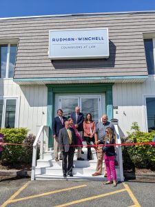 Group of men and women stand on the front steps of Rudman Winchell's Ellsworth office. Attorney Allison Economy, a woman with blonde hair, pink pants, and a navy blue shirt, prepares to cut a large red ribbon with over-sized ceremony scissors while others watch.