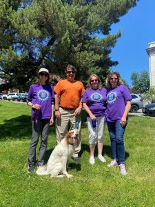 Group of 4 people and 1 dog posing for a picture outside on a sunny day. 3 people are wearing matching purple shirts supporting the Alzheimer's Association, and one person is wearing an orange shirt.