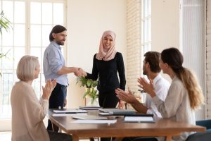 Group of colleagues sit and clap as new executive leader is welcomed to the group. Leader is an asian woman wearing a hijab. 