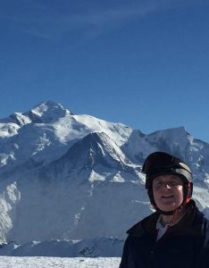 Justin Bennett stands in the bottom right corner of the photo wearing a black jacket, grey polo shirt, and a black helmet. Most of the photo is taken up by a snowy mountain and a clear blue sky. 