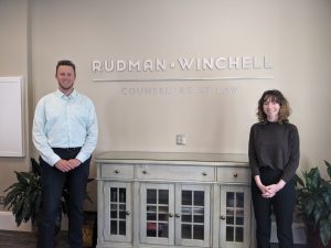 Ben and Bonnie stand in front of Rudman Winchell's logo in a conference room. 