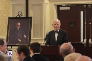 Hon. Paul L. Rudman Speaks in front of a crowd. He stands next to a painting of his brother, Gerald Rudman. 